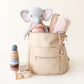 A beige diaper bag holds Eloise the Elephant - Little 13" by Cuddle + Kind, a pink blanket, and a pacifier; beside it are wooden blocks and a pastel stacking toy—making it an adorable birthday gift memory.