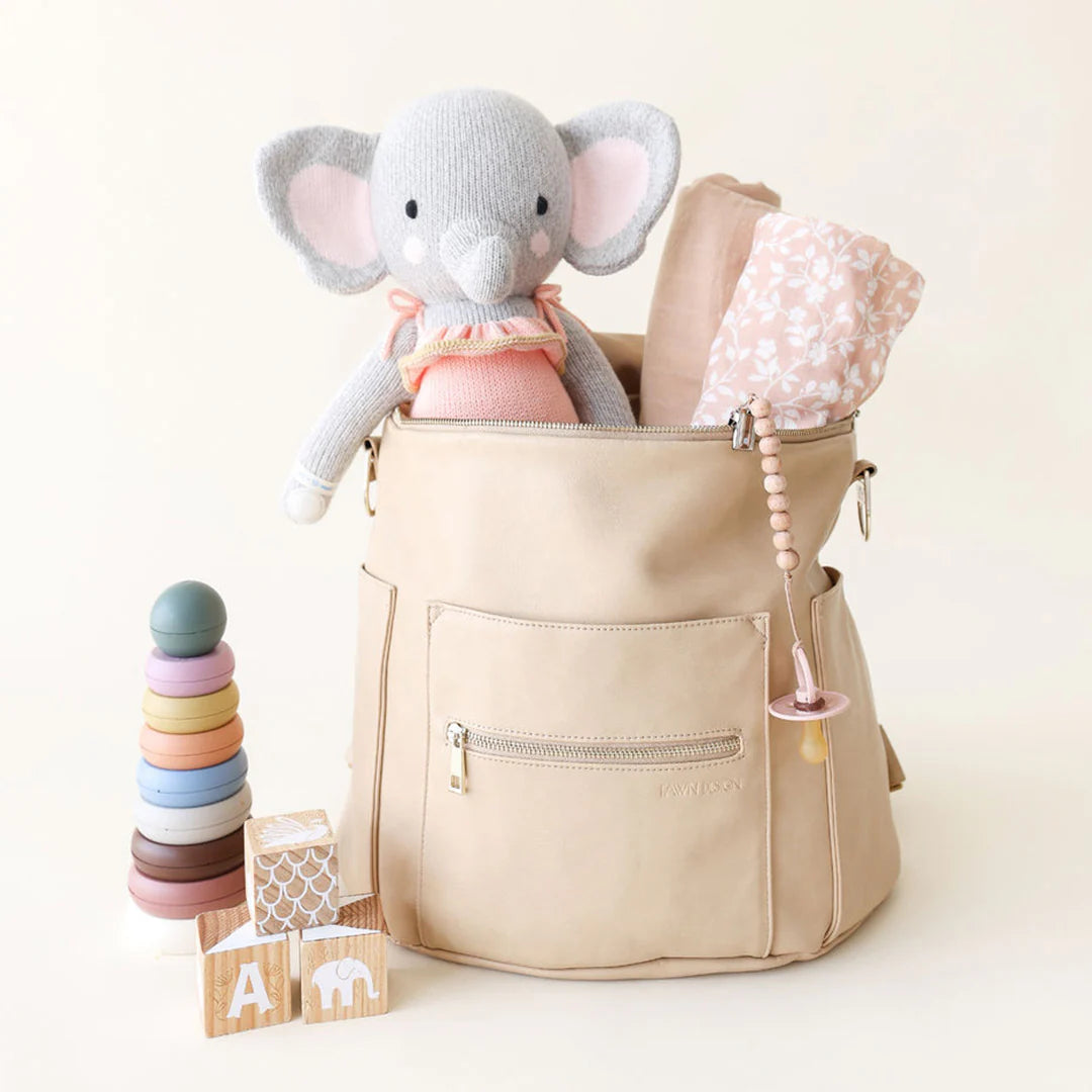 A beige diaper bag holds Eloise the Elephant - Little 13" by Cuddle + Kind, a pink blanket, and a pacifier; beside it are wooden blocks and a pastel stacking toy—making it an adorable birthday gift memory.