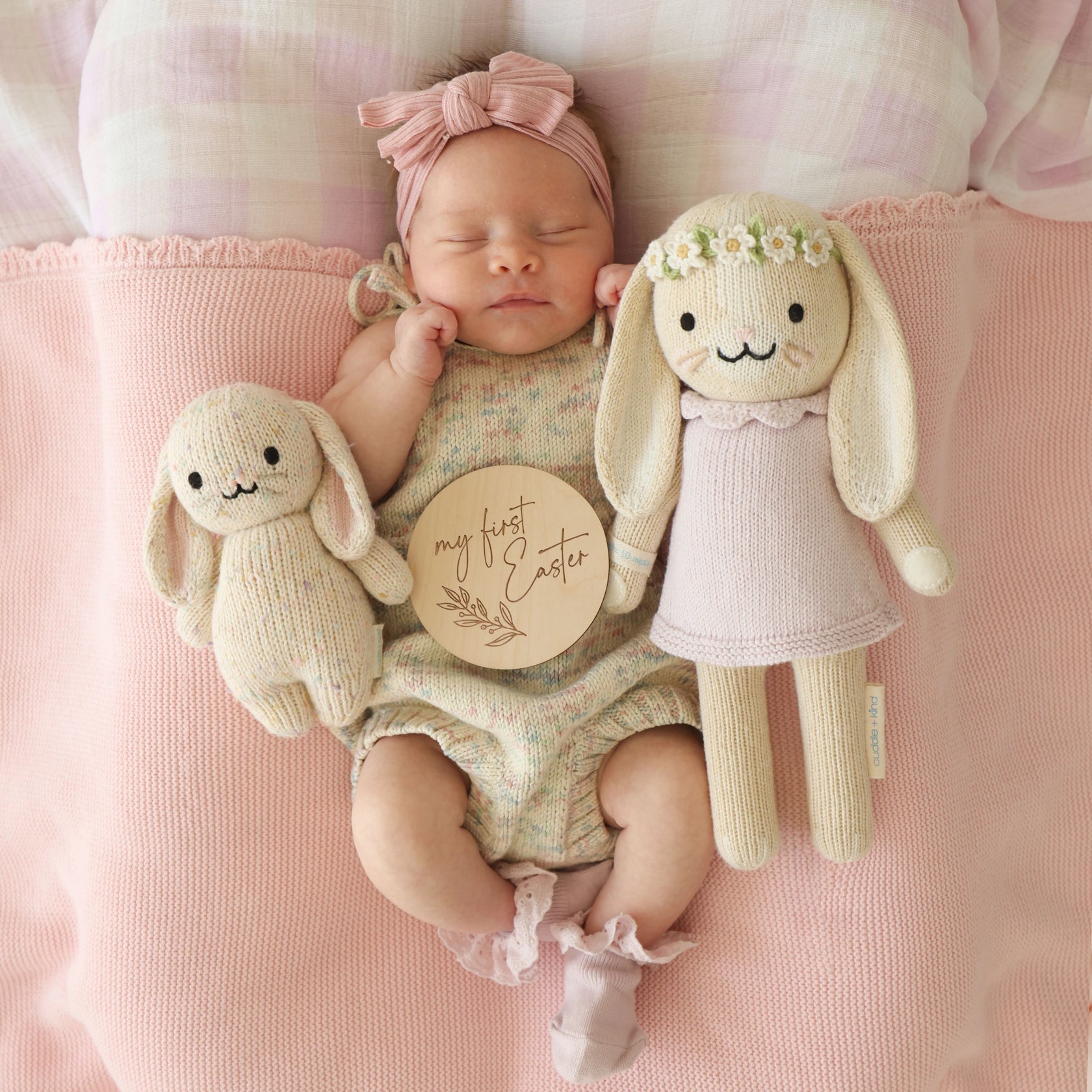 A newborn baby in a pink headband lies on a pink blanket, cuddling two Cuddle + Kind Hannah the Bunny Violet - Little 13" plush toys and holding a wooden plaque that reads "my first Easter"—a truly inspirational gift idea for welcoming little ones into the world.