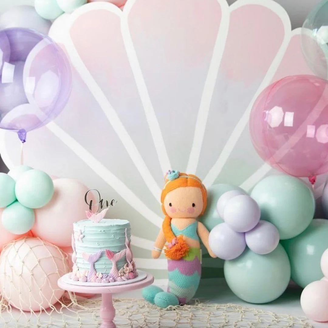 Step into a magical world at IslaBirthday with a pastel mermaid setup, featuring a shell backdrop, balloons, the Cuddle + Kind Isla the Mermaid - Little 13" doll, and a light blue cake topped with "one" and seashells.