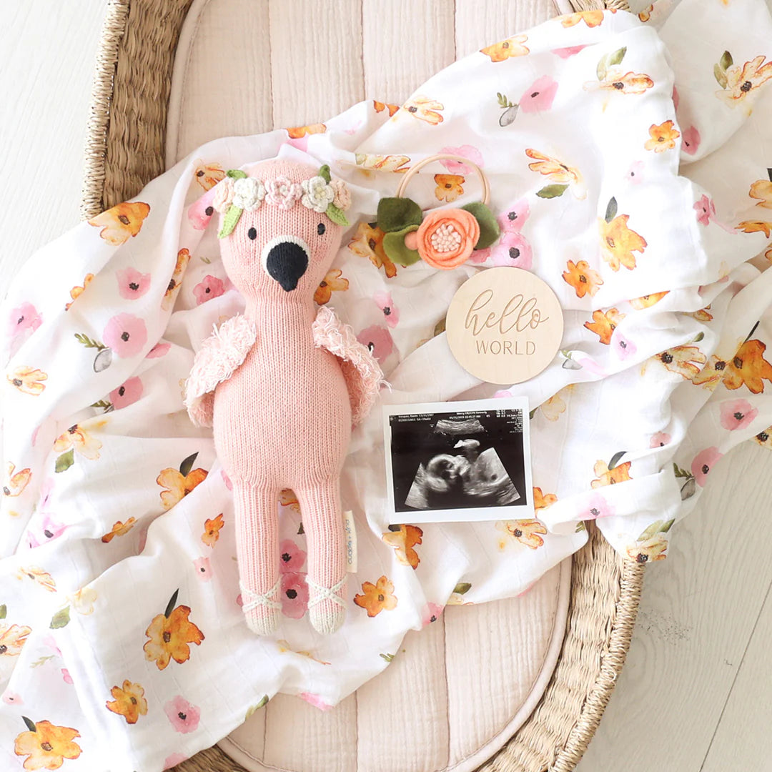 A Penelope the Flamingo by Cuddle + Kind, a floral blanket with tropical flowers, a wood "Hello World" sign, a flower hair clip, and an ultrasound photo are arranged in a wicker basket.