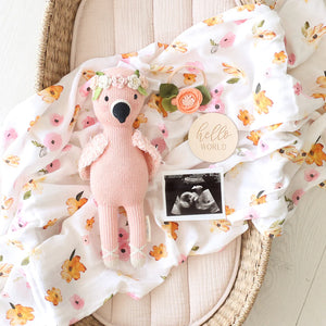 A Penelope the Flamingo by Cuddle + Kind, a floral blanket with tropical flowers, a wood "Hello World" sign, a flower hair clip, and an ultrasound photo are arranged in a wicker basket.