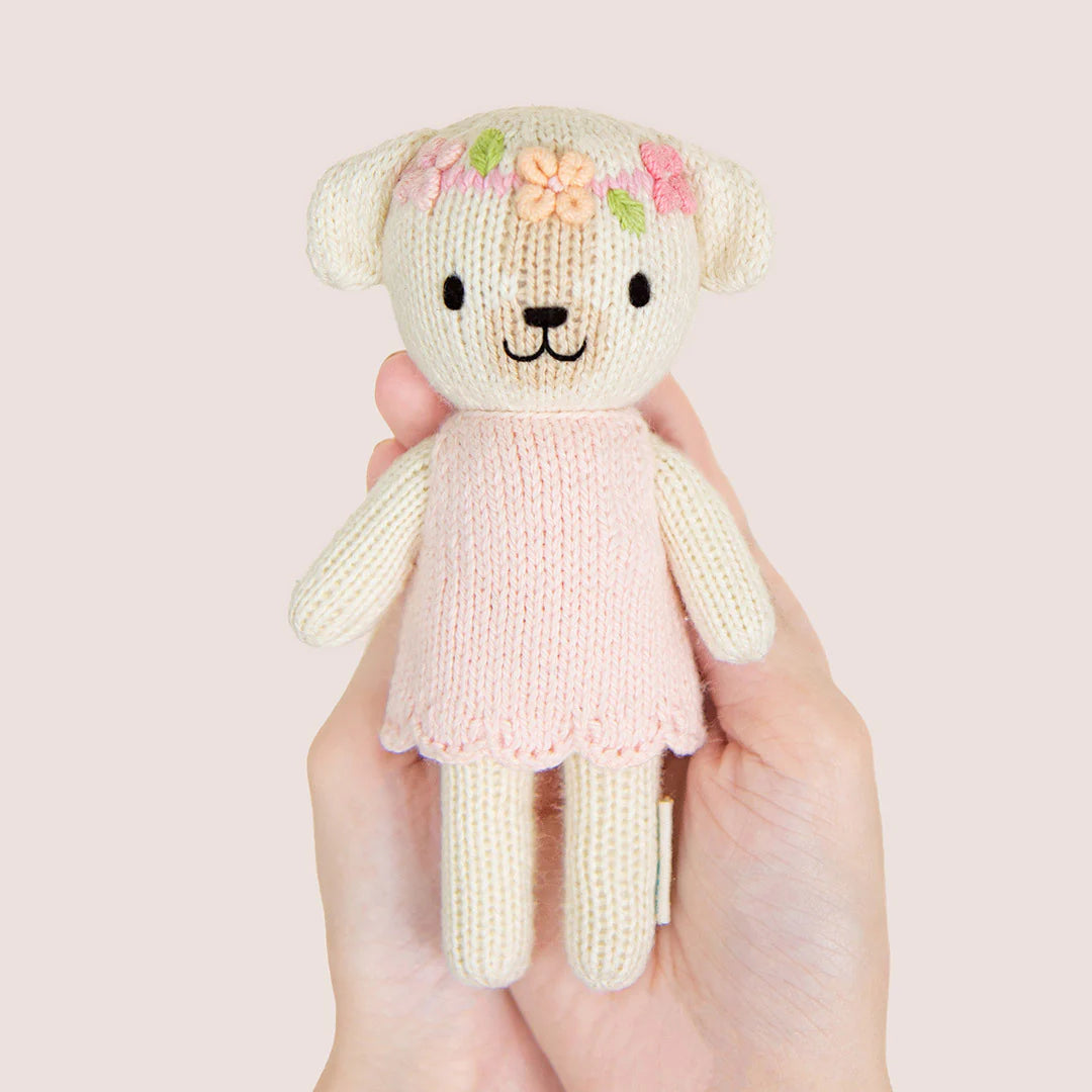 A hand is holding a Cuddle + Kind Tiny Charlotte the Dog, a fair trade hand-knit doll dressed in a pink dress with flower embroidery on its head.