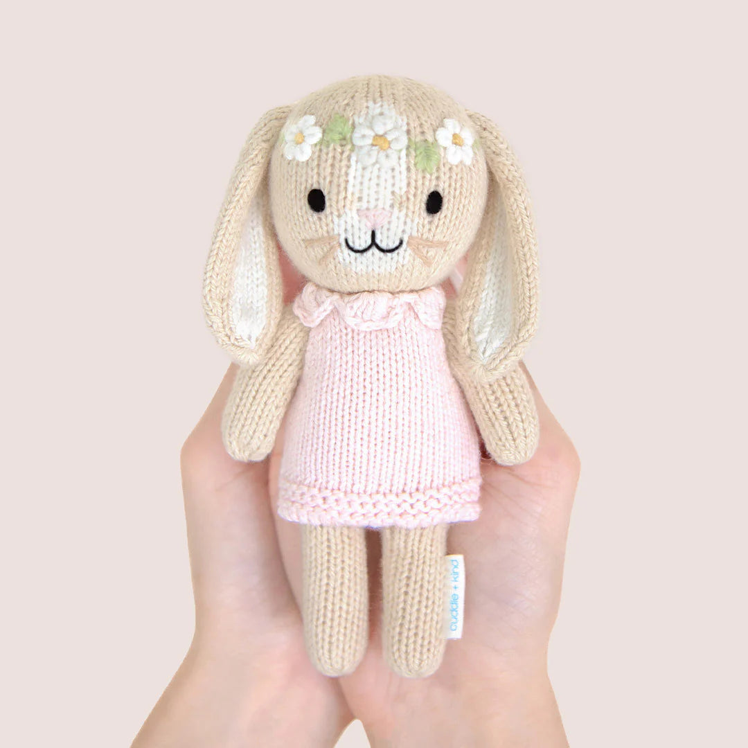 The Tiny Hannah the Bunny Blush doll by Cuddle + Kind, hand-knit and wearing a pink dress with flower accents, is held upright in two hands against a plain light background.