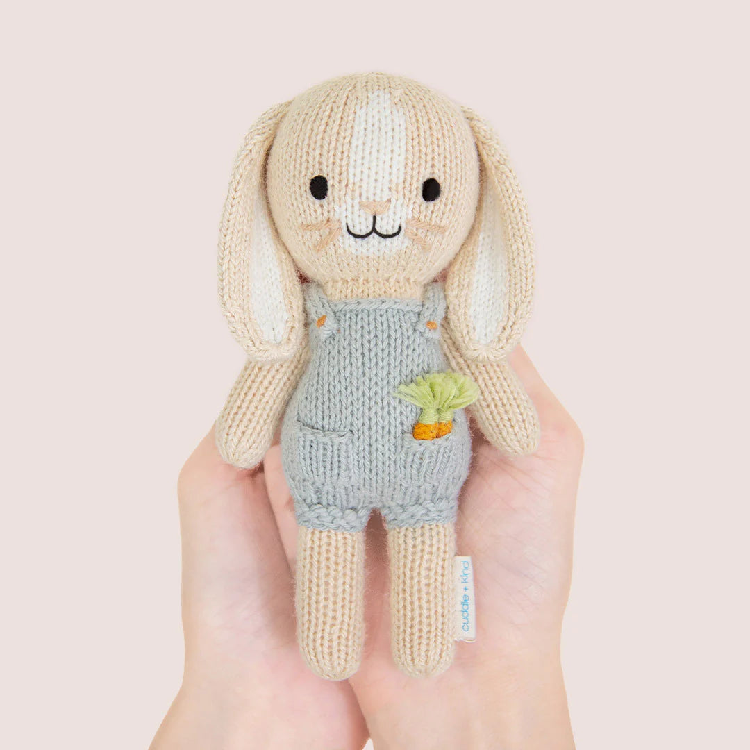 A hand holds Tiny Henry the Bunny, a hand-knit doll in light blue overalls with a carrot patch, from the Cuddle + Kind Tiny Collection.