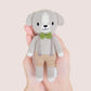 A hand holds Tiny Noah the Dog, a hand-knit doll from Cuddle + Kind's Tiny Collection, dressed in tan pants, gray suspenders, and a green bow tie, shown against a light background.