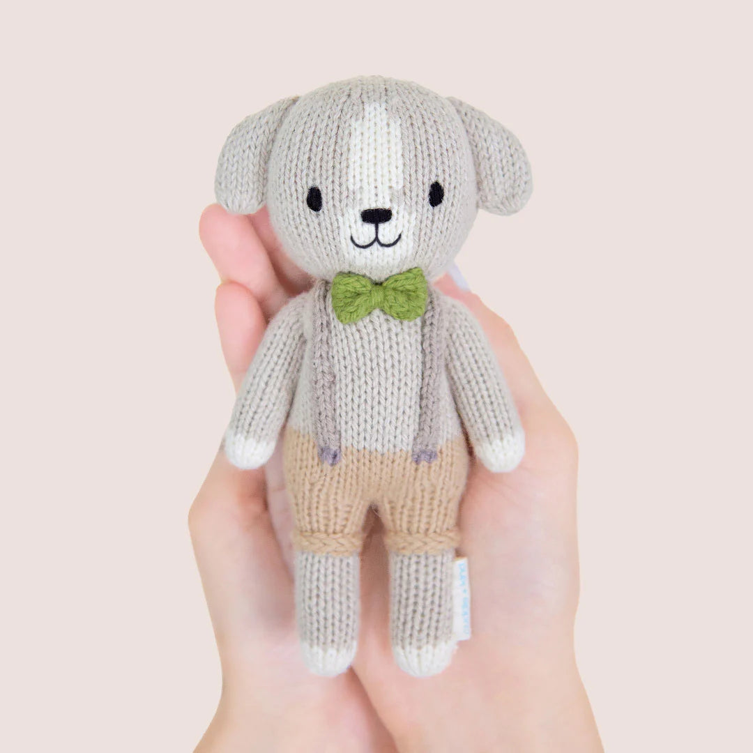 A hand holds Tiny Noah the Dog, a hand-knit doll from Cuddle + Kind's Tiny Collection, dressed in tan pants, gray suspenders, and a green bow tie, shown against a light background.