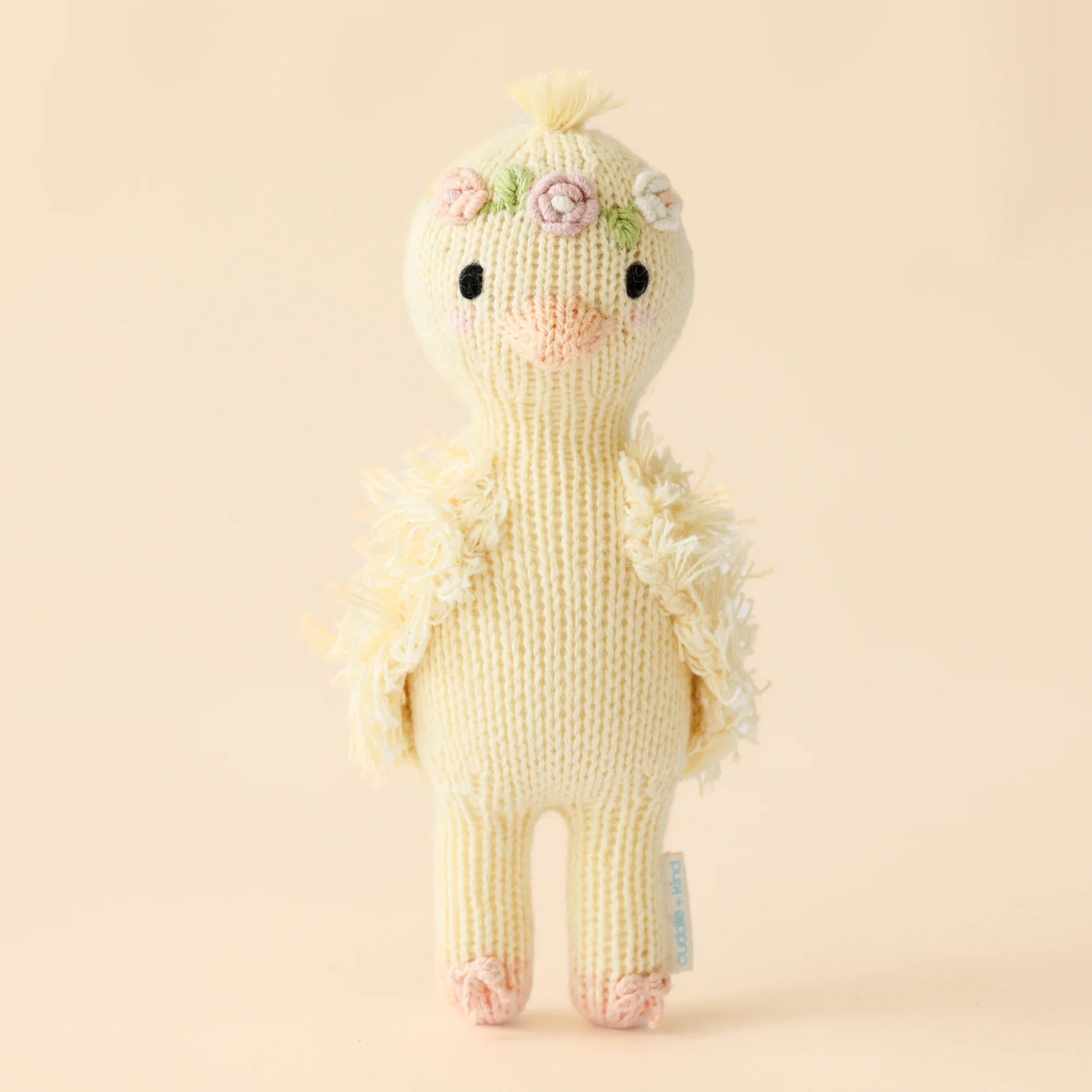 Tiny Flora the Duckling Mauve by Cuddle + Kind stands upright with fluffy wings, a flower crown, and pink feet—an artisan-made, fair trade plush from the Tiny Collection, shown against a plain beige background.