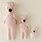 Three Cuddle + Kind Baby Flamingo dolls, hand-knit from heirloom Peruvian cotton yarn in pink and three sizes, are lined up on a neutral background—two wear flower crowns, one has a fluffy head.
