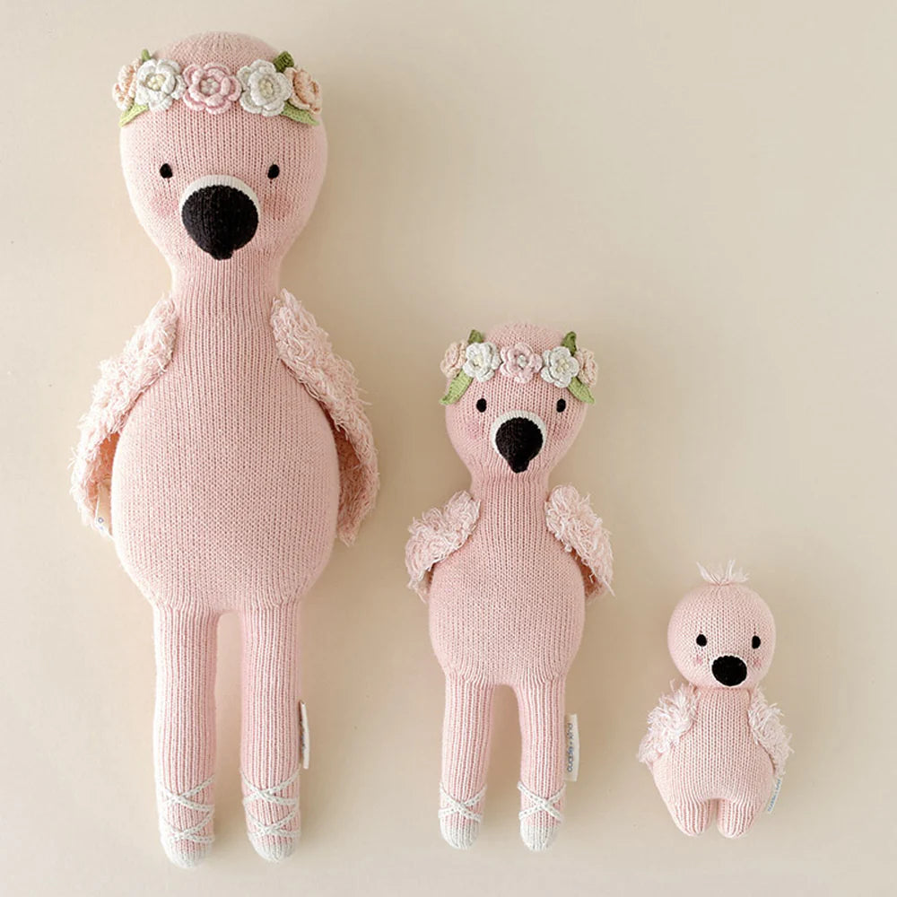 Three Cuddle + Kind Baby Flamingo dolls, hand-knit from heirloom Peruvian cotton yarn in pink and three sizes, are lined up on a neutral background—two wear flower crowns, one has a fluffy head.
