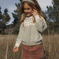 A young girl with long hair stands in a grassy field on a sunny day, wearing the Rylee & Cru Girls’ Padded Headband along with a patterned sweater and corduroy skirt, with trees visible in the background.