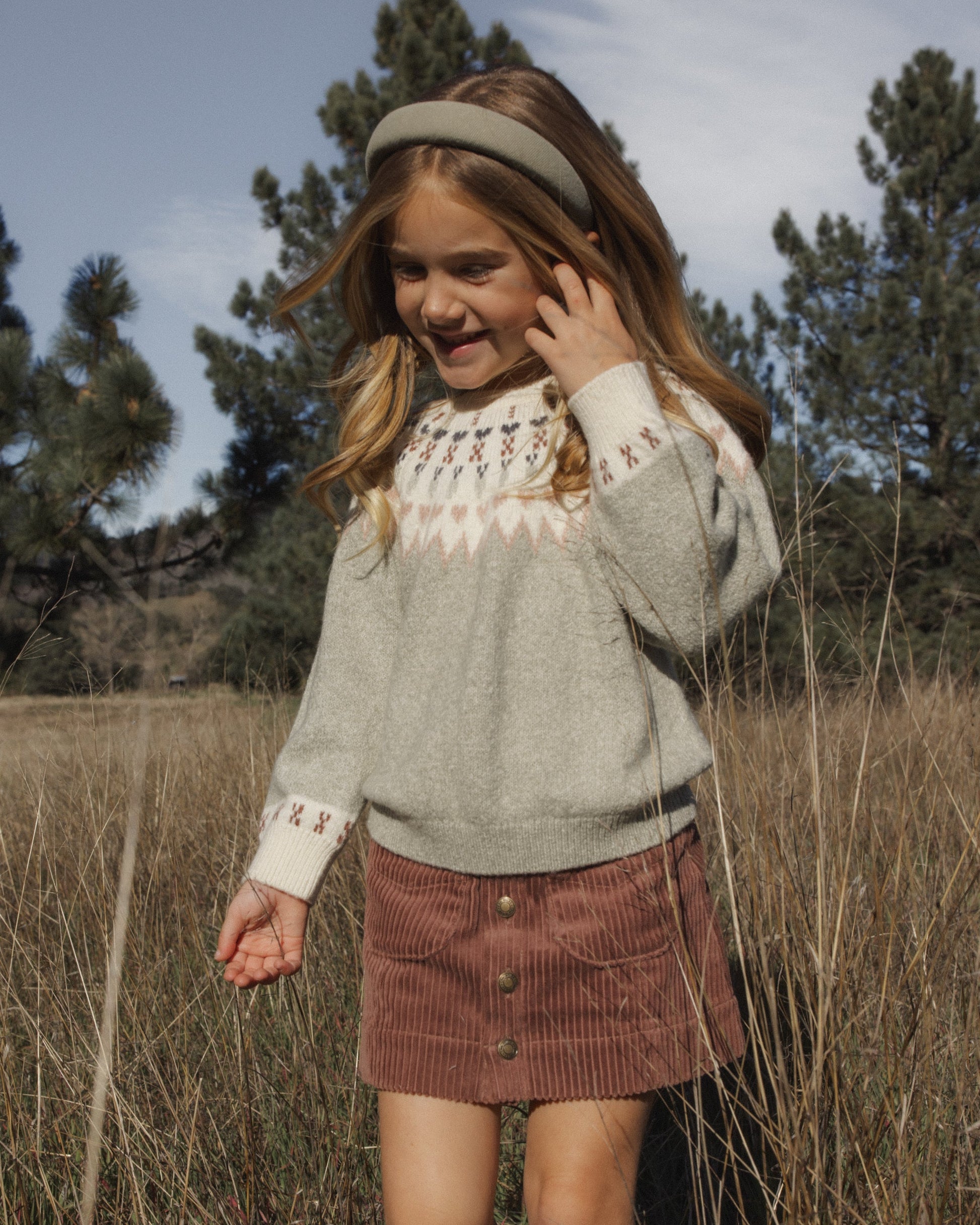 A young girl with long hair stands in a grassy field on a sunny day, wearing the Rylee & Cru Girls’ Padded Headband along with a patterned sweater and corduroy skirt, with trees visible in the background.