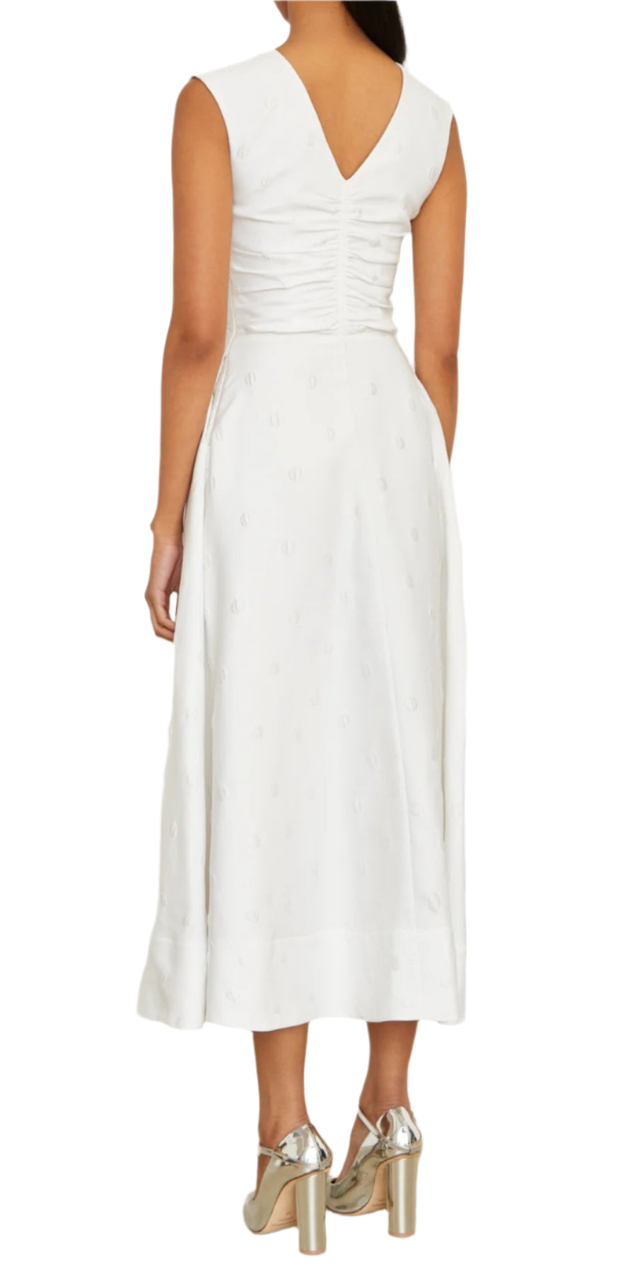 A woman in the Eliza Dress by Brandon Maxwell, featuring a white, sleeveless, V-neck midi design crafted from an Italian silk blend with subtle patterns, stands facing away. She completes her look with silver high heels, embodying essential resort-season elegance.
