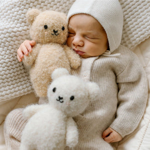 A baby in a cream knit outfit and hat, crafted from heirloom-quality Peruvian cotton yarn, sleeps on a textured blanket, holding a beige teddy bear with the Cuddle + Kind Ivory Boucle Bear - Baby 7" nearby.