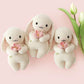Three Cuddle + Kind “Baby Bunny - Blooming Tulip” plush toys, lovingly hand-knit from cotton yarn in a soft cream hue and each holding a pink flower, are arranged beside two white tulips on a pale pink background—a charming heirloom-quality collection of baby animal companions.