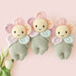 Three Cuddle + Kind Baby Daisy – Sunshower knitted plush dolls with pastel flower faces and green bodies are arranged side by side on a light pink background next to two white tulips—each one a soft baby companion perfect for snuggles and treasured moments.
