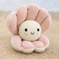Baby Pearl by Cuddle + Kind, a handcrafted plush toy made from natural Peruvian cotton and shaped like a pink seashell with a smiling face, sits on sandy beach with blurred water in the background.