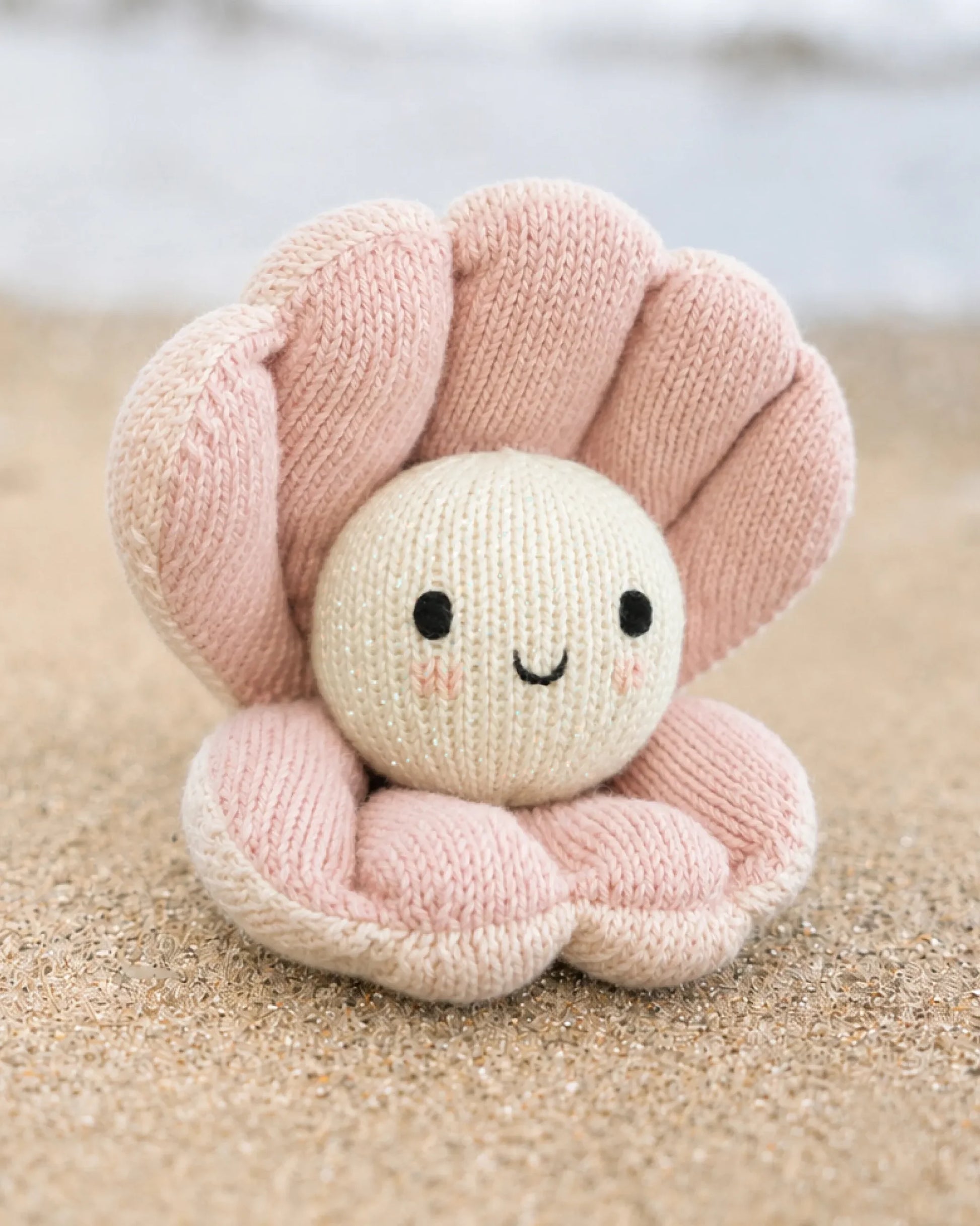 Baby Pearl by Cuddle + Kind, a handcrafted plush toy made from natural Peruvian cotton and shaped like a pink seashell with a smiling face, sits on sandy beach with blurred water in the background.