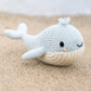 The Cuddle + Kind Baby Whale, a handcrafted plush toy knit from heirloom-quality Peruvian cotton in cheerful blue and white, smiles gently as it rests on a sandy beach with blurred water behind—an irresistibly soft companion for babies.