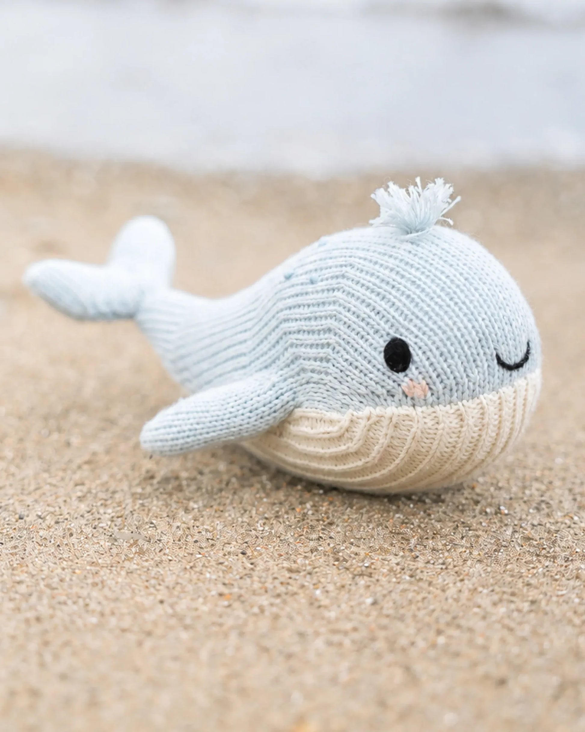 The Cuddle + Kind Baby Whale, a handcrafted plush toy knit from heirloom-quality Peruvian cotton in cheerful blue and white, smiles gently as it rests on a sandy beach with blurred water behind—an irresistibly soft companion for babies.