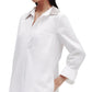 A person wears the TWP Jennys Gown with a crystal collar, crafted from Italian cotton shirting. Their left hand is in a pocket, and their right arm is slightly bent.