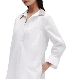 A person wears the TWP Jennys Gown with a crystal collar, crafted from Italian cotton shirting. Their left hand is in a pocket, and their right arm is slightly bent.