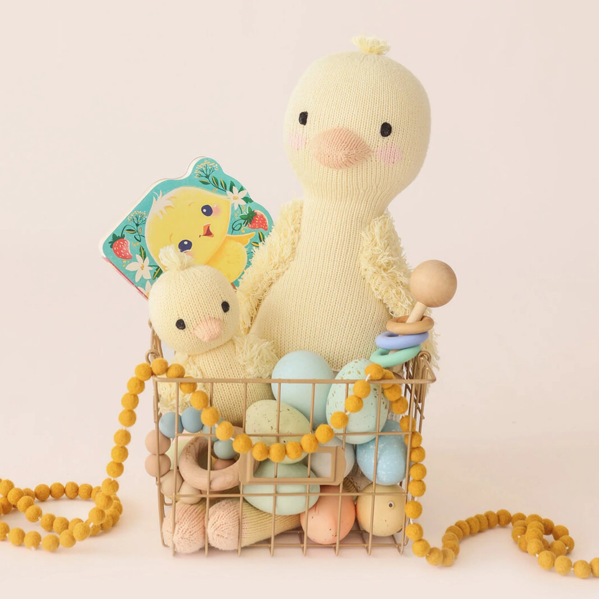Cuddle + Kind’s Finley the Duckling 20" plush sits in a wire basket with pastel eggs, a baby rattle, duck book, and yellow pom-pom garland—an adorable birthday gift arrangement on a light background.