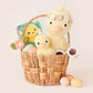 A woven basket holds pastel eggs, Cuddle + Kind’s Flora the Duckling, Mauve - Regular 20", a smaller duck plush, a book, felt wildflowers, and white sunglasses—perfect for an afternoon by a sparkling river.