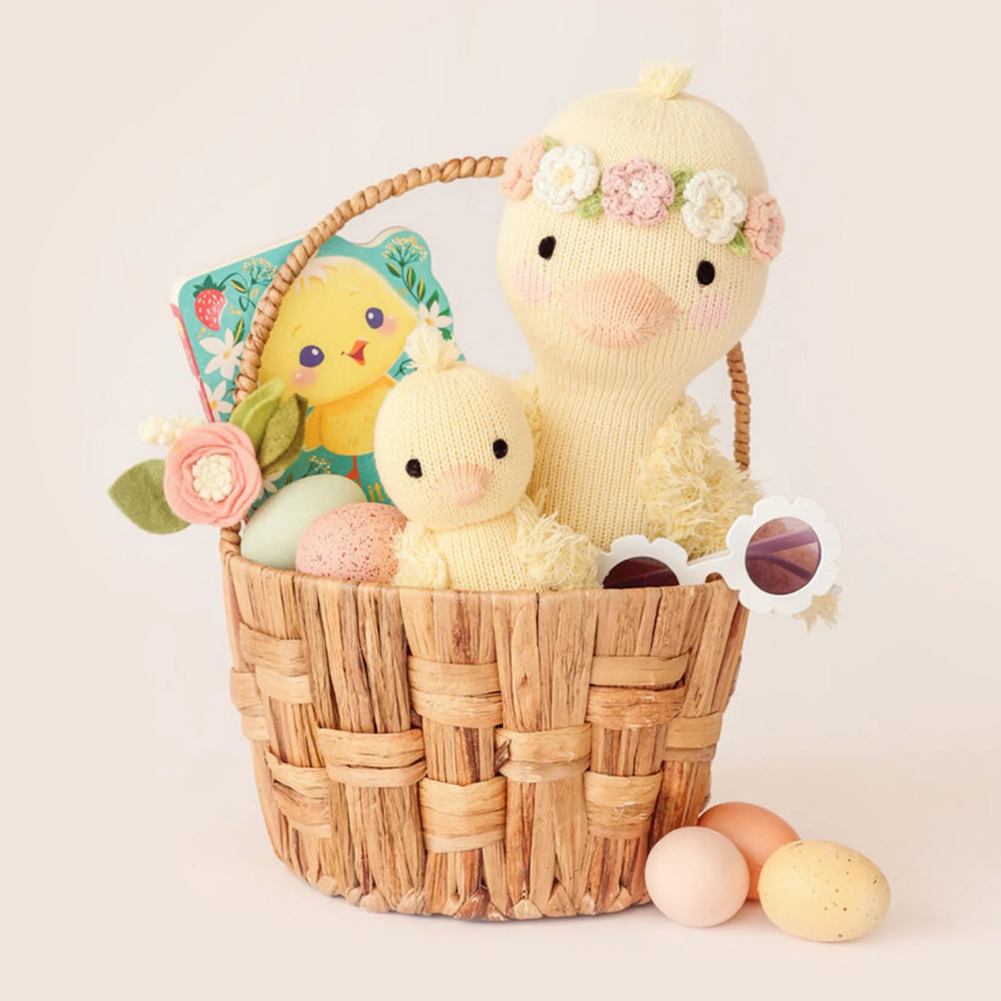 A woven basket featuring two Cuddle + Kind Flora the Duckling Mauve - Little 13" plush toys, pastel eggs, a book, toy sunglasses, and felt flowers, with three pastel eggs placed beside the basket.