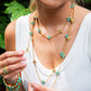 A woman in a white sleeveless top wears the Juliska Capucine De Wulf Berry Clover Double Sided Necklace, featuring layered gold and turquoise details, and holds one necklace with her hand.