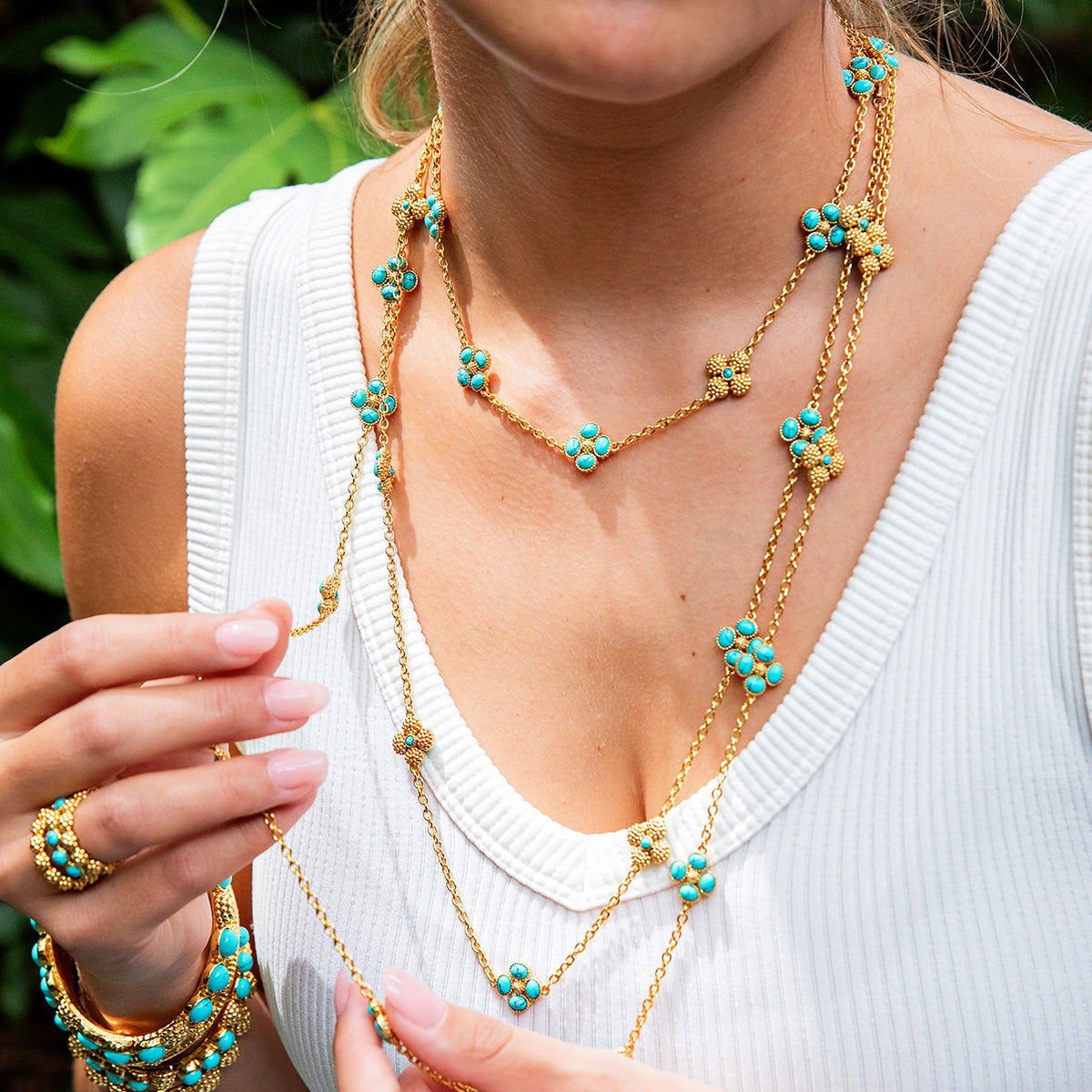 A woman in a white sleeveless top wears the Juliska Capucine De Wulf Berry Clover Double Sided Necklace, featuring layered gold and turquoise details, and holds one necklace with her hand.