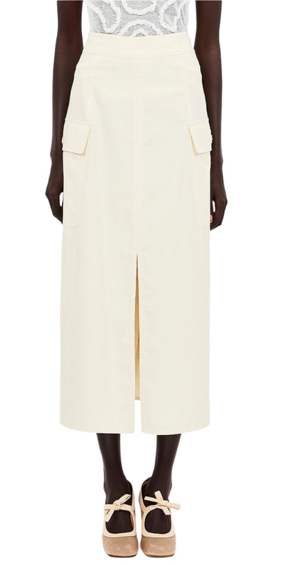 An individual is dressed in a white patterned top and an Ulla Johnson Rhodes Skirt, a cream-colored cotton tencel piece with pockets and front slit. They complement it with beige platform shoes featuring ankle straps.