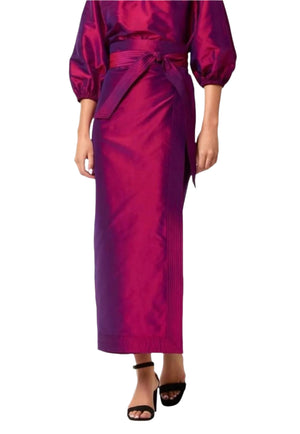 A person wearing the Ann Mashburn Kira Silk Shantung Wrap Skirt in magenta with a matching belt, a puff-sleeve top, and black open-toe heels.