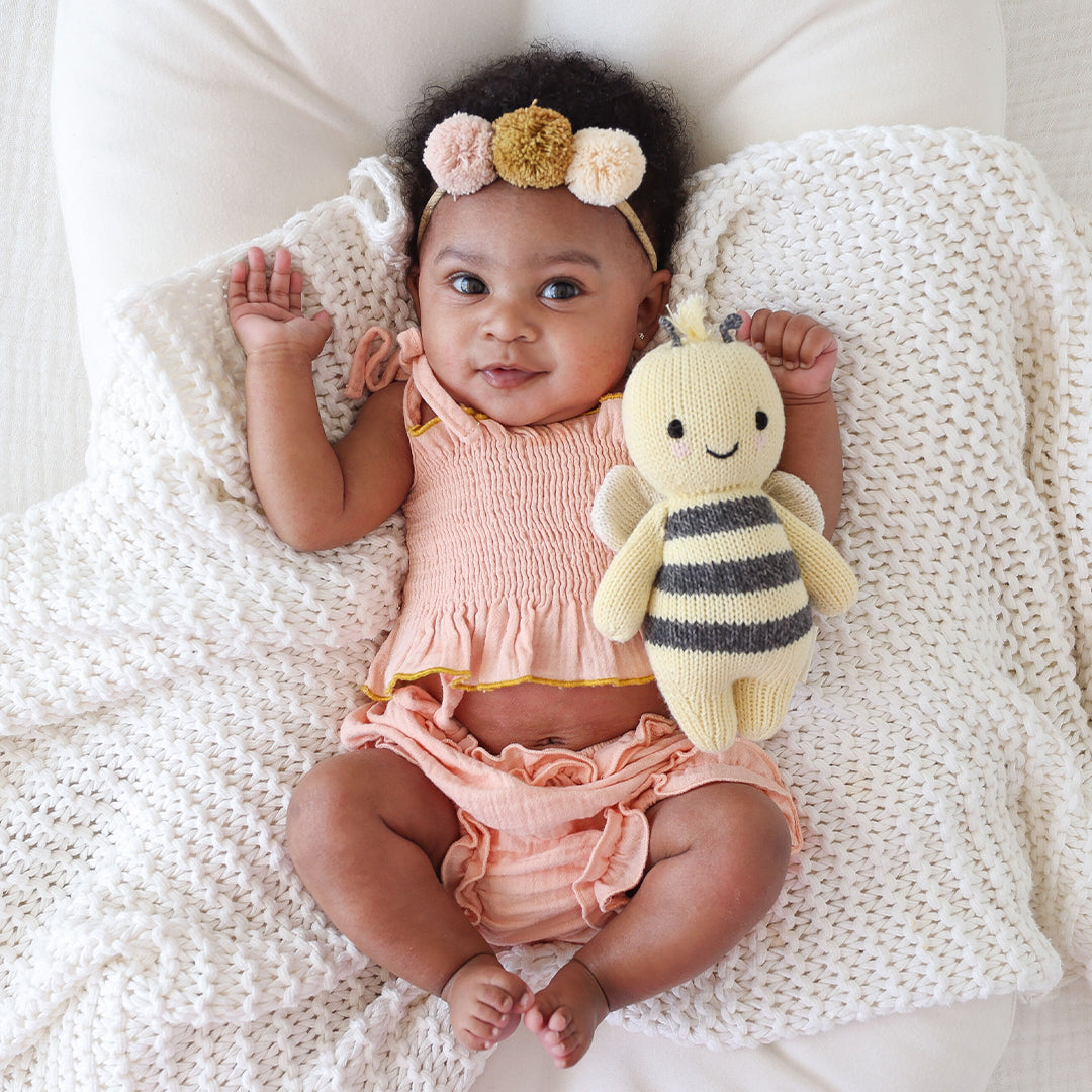 A baby in a peach outfit lies on a white textured blanket, holding the Cuddle + Kind Bee – Baby 7” heirloom-quality, hand-knit toy and wearing a pom-pom headband.