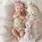 A baby wearing a pink headband and knit romper lies on a textured blanket next to three heirloom-quality, hand-knit baby animals—including the Cuddle + Kind Strawberry Cow - Baby 7"—crafted from soft Peruvian cotton yarn.