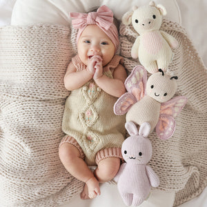 A baby wearing a pink headband and knit romper lies on a textured blanket next to three heirloom-quality, hand-knit baby animals—including the Cuddle + Kind Strawberry Cow - Baby 7"—crafted from soft Peruvian cotton yarn.