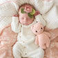 A newborn baby sleeps on a patterned blanket, wearing a flower headband and holding the "Piglet - Baby 7"" by Cuddle + Kind, a pink hand-knit baby animal crafted from heirloom-quality Peruvian cotton yarn.