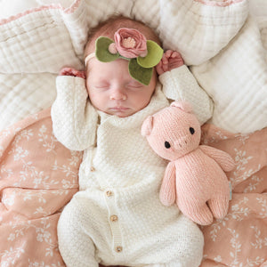 A newborn baby sleeps on a patterned blanket, wearing a flower headband and holding the "Piglet - Baby 7"" by Cuddle + Kind, a pink hand-knit baby animal crafted from heirloom-quality Peruvian cotton yarn.
