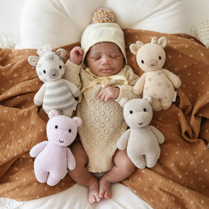 A baby wearing a cream outfit and hat lies on a brown blanket, surrounded by four heirloom-quality, hand-knit baby animals—including the Cuddle + Kind Zebra - Baby 7"—in white, beige, gray, and light pink; each one crafted from soft Peruvian cotton yarn.