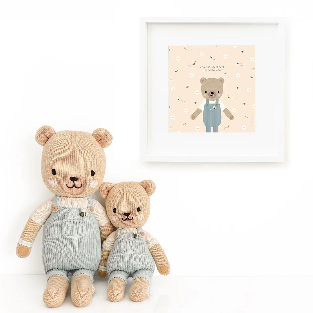 Two Charlie the Honey Bear dolls by Cuddle + Kind, each 13", sit side by side in matching overalls before a white wall with a framed bear-in-overalls print on a floral background.