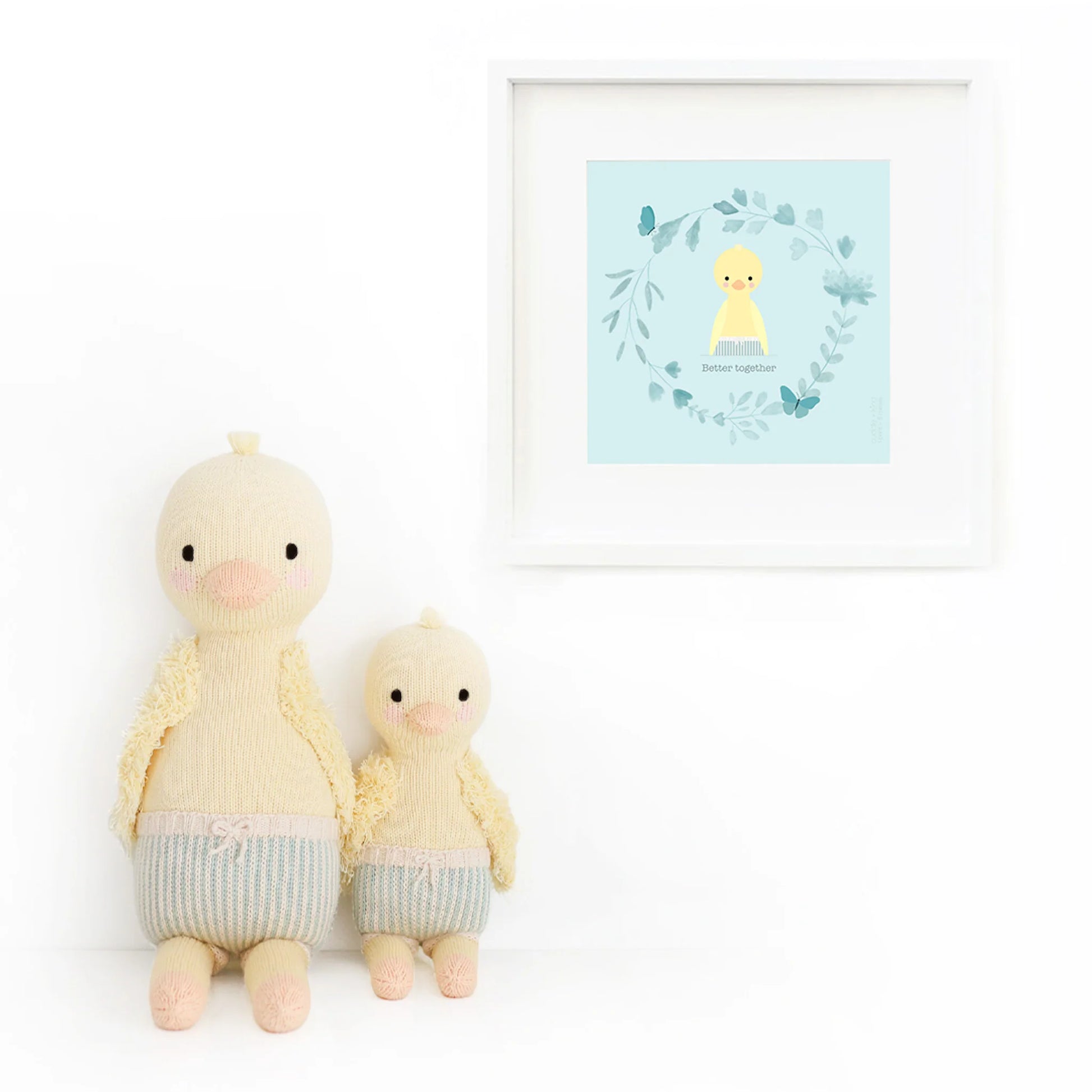 Finley the Duckling by Cuddle + Kind—a cuddly 20" plush—sits by a wall with a framed yellow duck and leaf wreath, creating an adorable gift scene perfect for any birthday or duck lover.