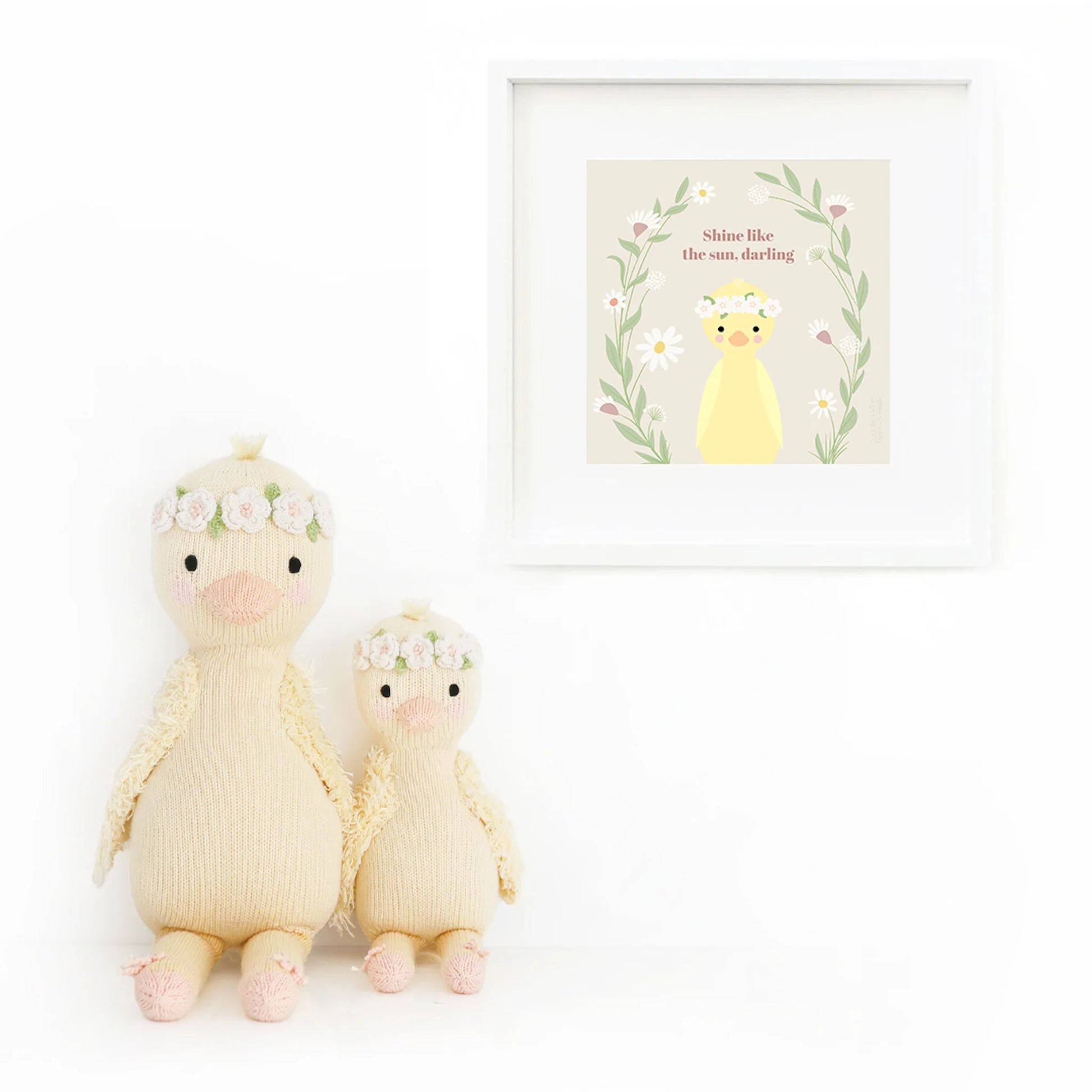 Two Flora the Duckling Ivory - Little 13" plush toys by Cuddle + Kind sit beside a wall art print with a yellow duck, wildflowers, and the phrase "Shine like the sun, darling," inspiring brilliance.