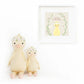 Two Cuddle + Kind Flora the Duckling Ivory plush toys sit beside a framed print with a yellow duck, wildflowers, and the words "Shine like the sun, darling," displayed on a white wall.