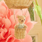 A Cuddle + Kind Tiny Charlie the Honey Bear, hand-knit and dressed in overalls, sits in a small wicker basket hanging from a hot air balloon with a large pink paper flower and a window in the background.