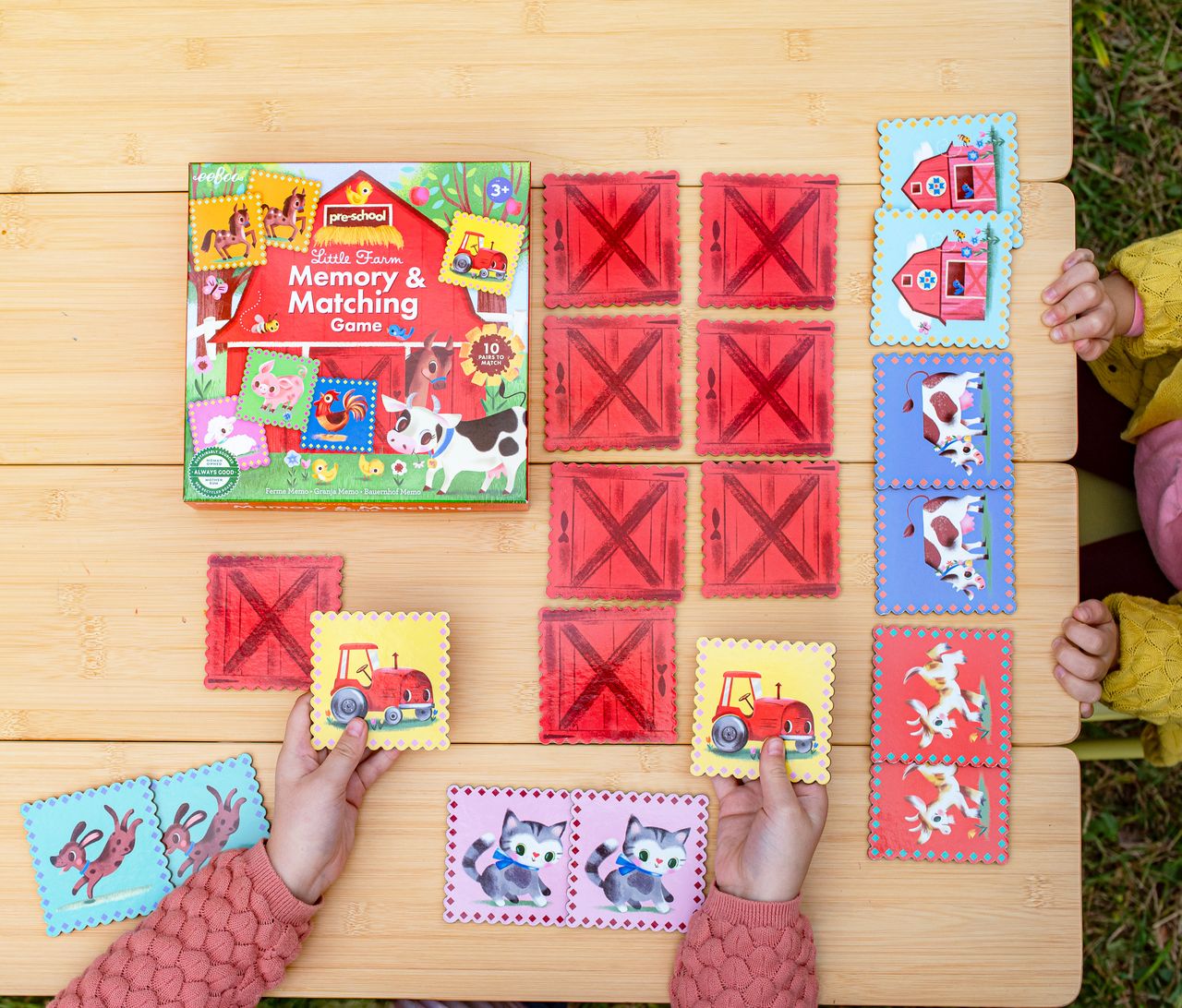 Two children play Eeboo’s Little Farm Pre-School Matching Game at a wooden table, challenging their minds with cards featuring animals and vehicles. The game’s box is also on the table, making this engaging memory game for kids a perfect choice for learning and play.