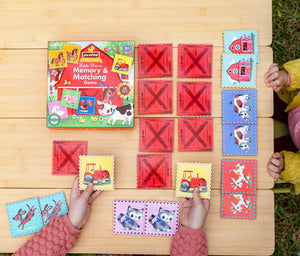 Two children play Eeboo’s Little Farm Pre-School Matching Game at a wooden table, challenging their minds with cards featuring animals and vehicles. The game’s box is also on the table, making this engaging memory game for kids a perfect choice for learning and play.
