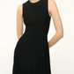 A person wearing the Staud Elyse Dress—a sleeveless black ribbed knit dress with a crew neckline, fitted bodice, and pleated skirt—stands against a plain background.