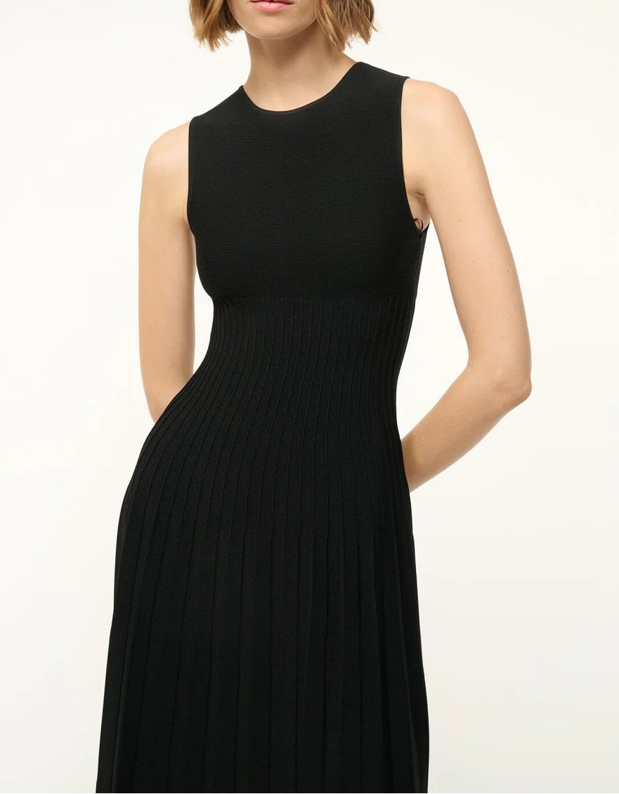A person wearing the Staud Elyse Dress—a sleeveless black ribbed knit dress with a crew neckline, fitted bodice, and pleated skirt—stands against a plain background.