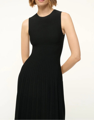 A person wearing the Staud Elyse Dress—a sleeveless black ribbed knit dress with a crew neckline, fitted bodice, and pleated skirt—stands against a plain background.