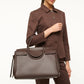 A woman in a brown suit holds the Staud Maude Carryall, an oversized bag crafted from structured dark brown pebbled leather, by its handles against a plain background.